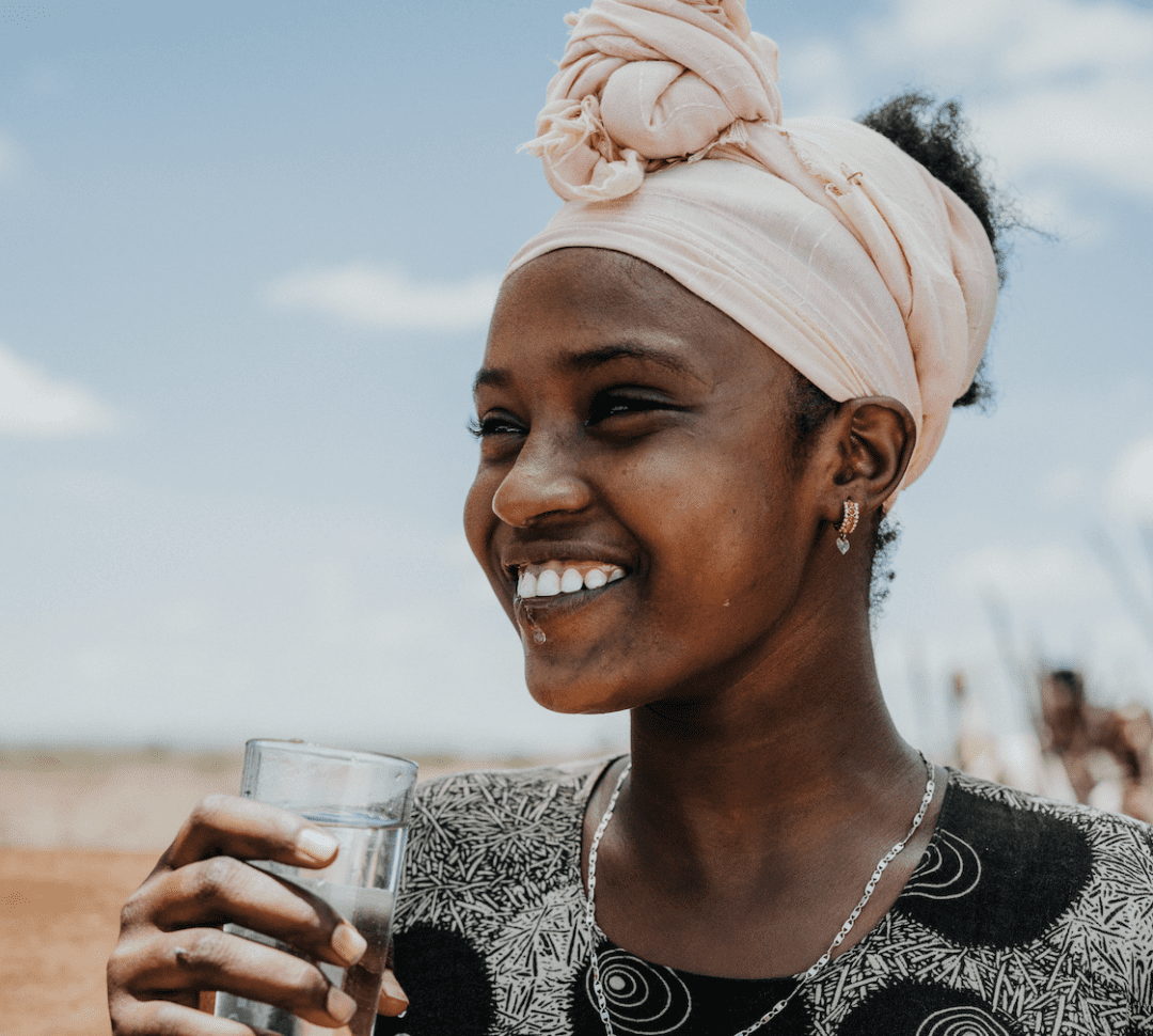 A smiling woman wearing a pale pink headwrap and patterned dress holds a glass of water outdoors on a sunny day, with blue sky and blurred landscape in the background.